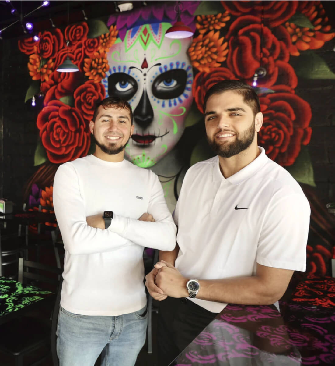 Two smiling men in white shirts stand inside a restaurant with a colorful Day of the Dead mural behind them.