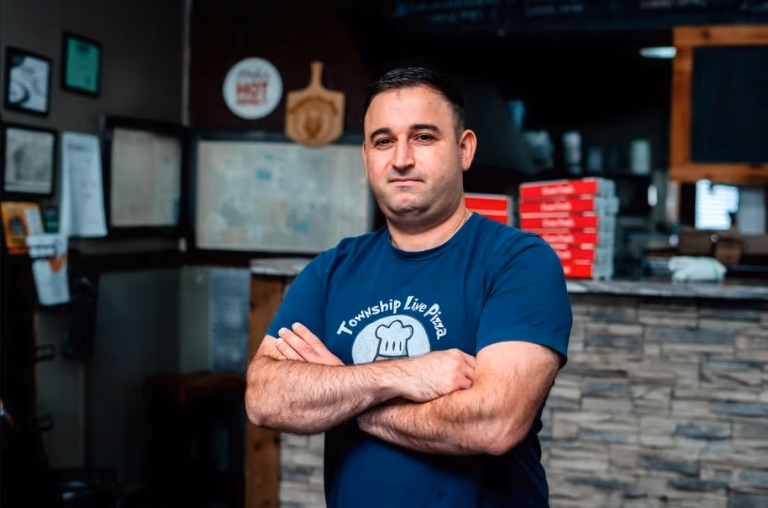 Man in a blue Township Live Pizza t-shirt stands with arms crossed in a pizza restaurant interior.