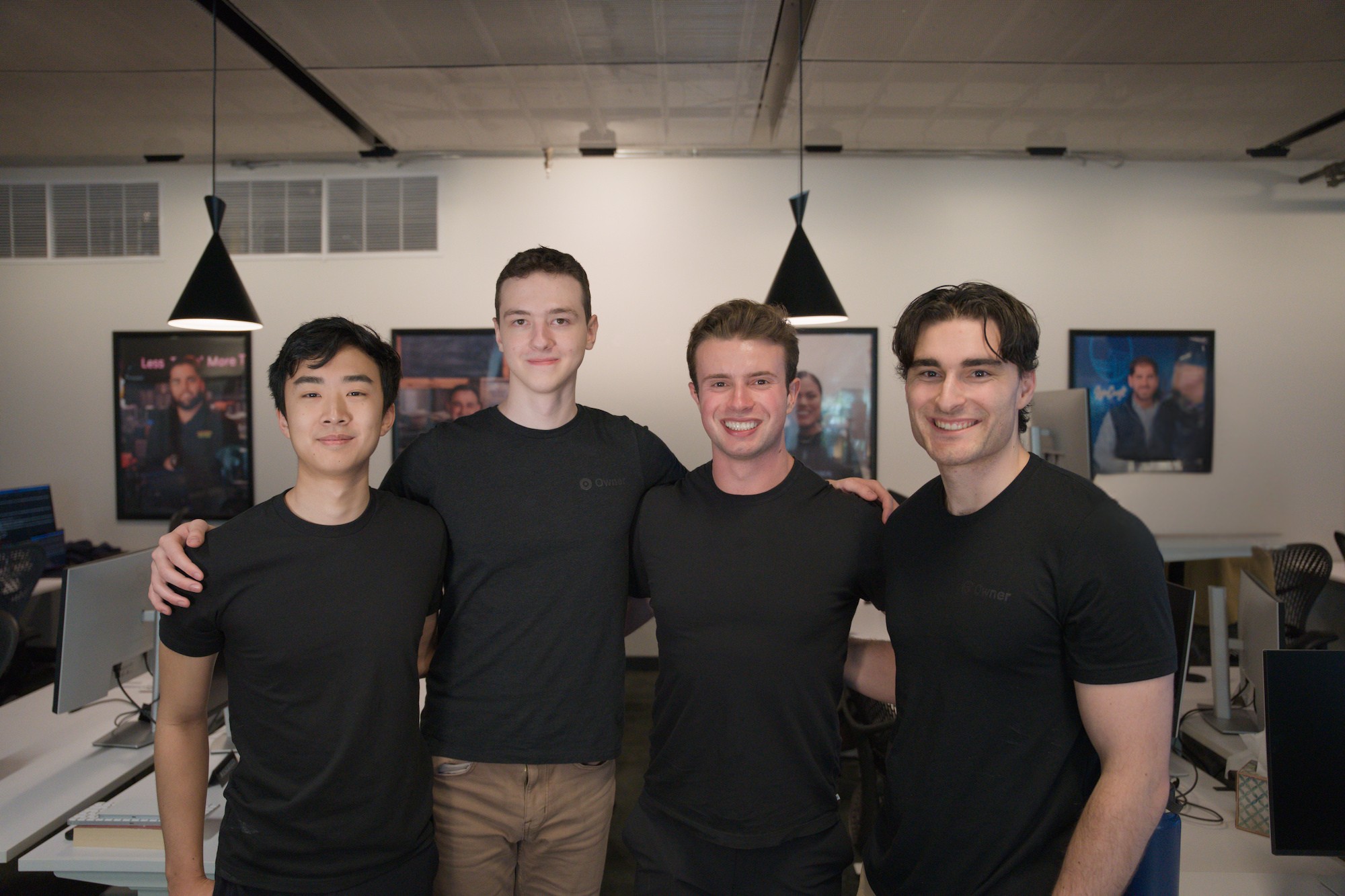 Four young men standing closely and smiling inside a modern office space.