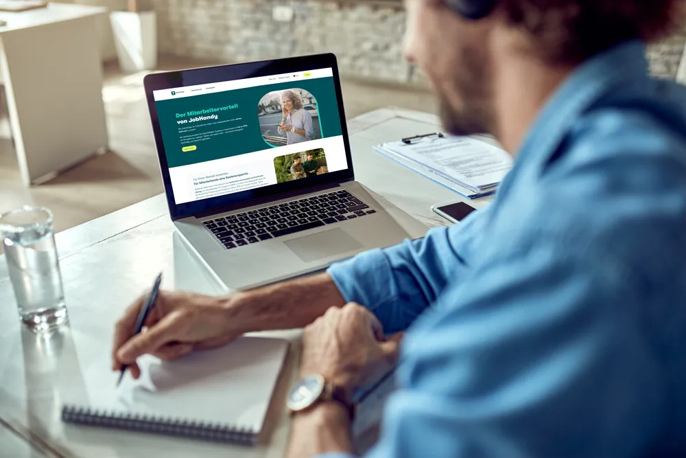 Mann arbeitet am Schreibtisch, schreibt in Notizbuch vor einem Laptop mit einer Webseite über Mitarbeiterangebote von JobHandy.
