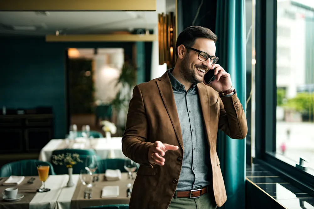 Ein lächelnder Mann mit Brille und brauner Jacke führt ein Telefonat in einem eleganten Restaurant bei Tageslicht.