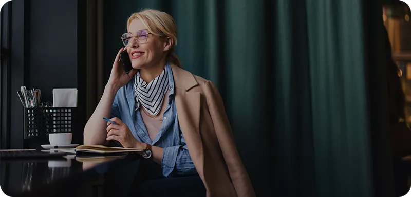 Frau mit Brille spricht lächelnd am Telefon, sitzt in einem Café und hält einen Stift über ein Notizbuch.