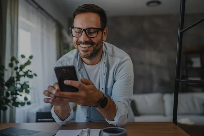 Lächelnder Mann mit Brille sitzt an einem Tisch und schaut auf sein Smartphone in einem hellen Raum.