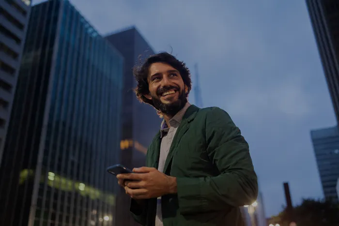 Ein lächelnder Mann mit dunkelgrüner Jacke hält ein Smartphone vor einer Stadtlandschaft bei Abenddämmerung.