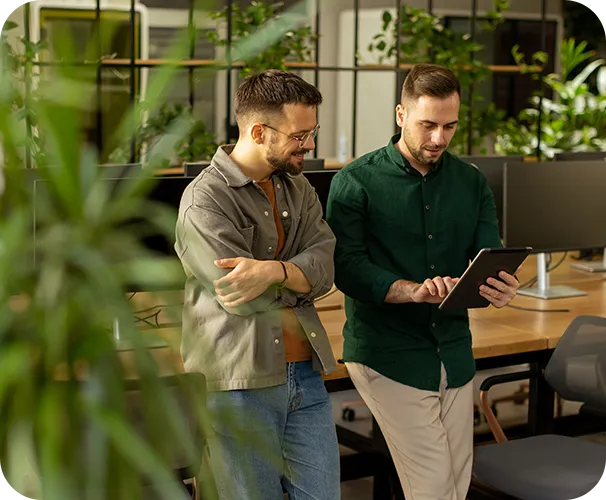 Zwei Männer in einem modernen Büro betrachten gemeinsam ein Tablet, umgeben von grünen Pflanzen.