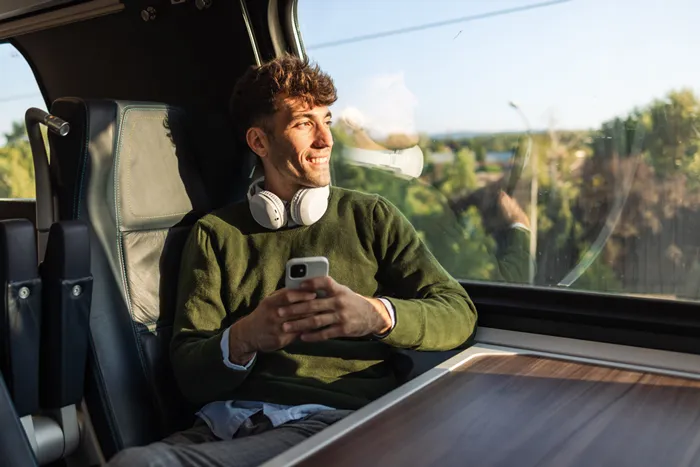 Junger Mann mit Kopfhörern um den Hals sitzt in einem Zug, hält ein Smartphone und schaut lächelnd aus dem Fenster.
