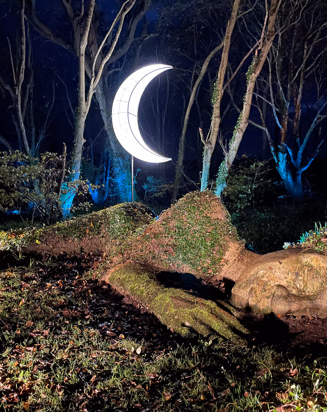 Large moss-covered sculpture of a reclining figure with a glowing crescent moon suspended above in a dark forest.