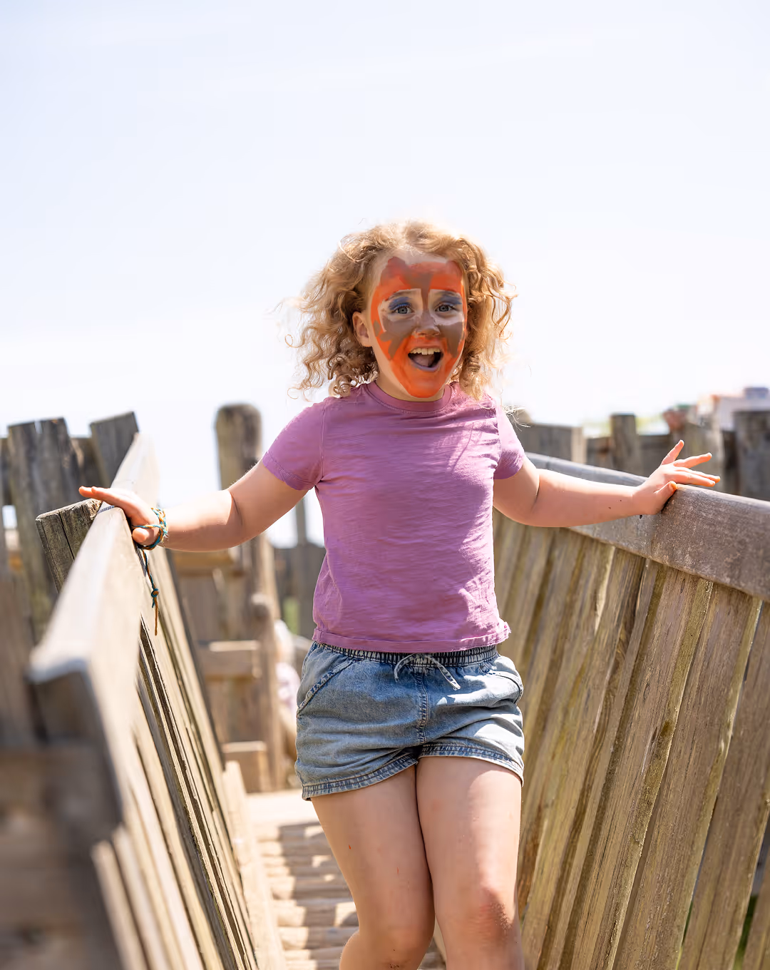 Smiling young girl with curly hair and face paint standing on a wooden bridge outdoors.