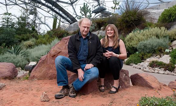 Blonde woman wearing a blue jacket standing outdoors near pink flowering bushes.