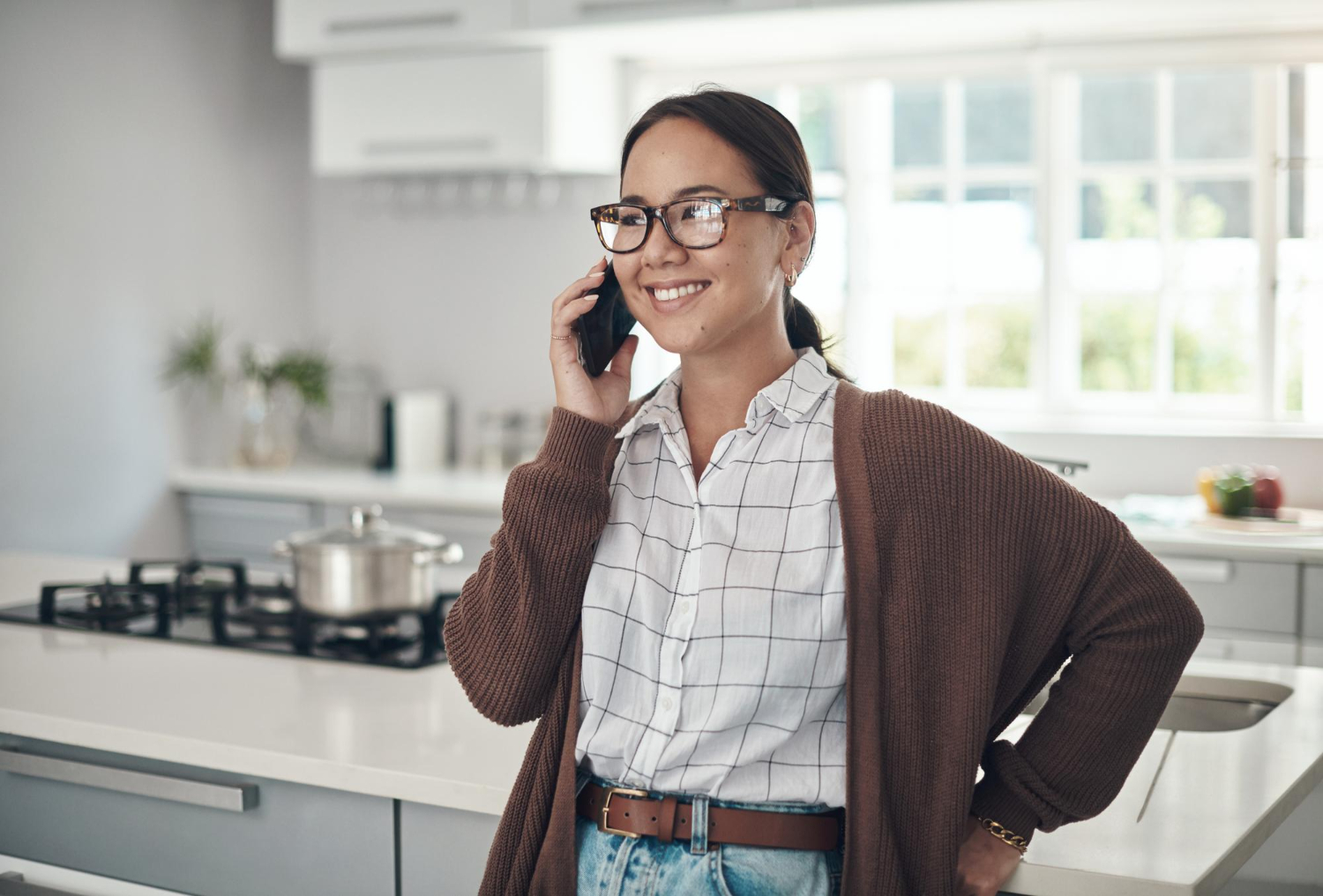 Smiling woman with glasses talking on a smartphone in a bright kitchen.