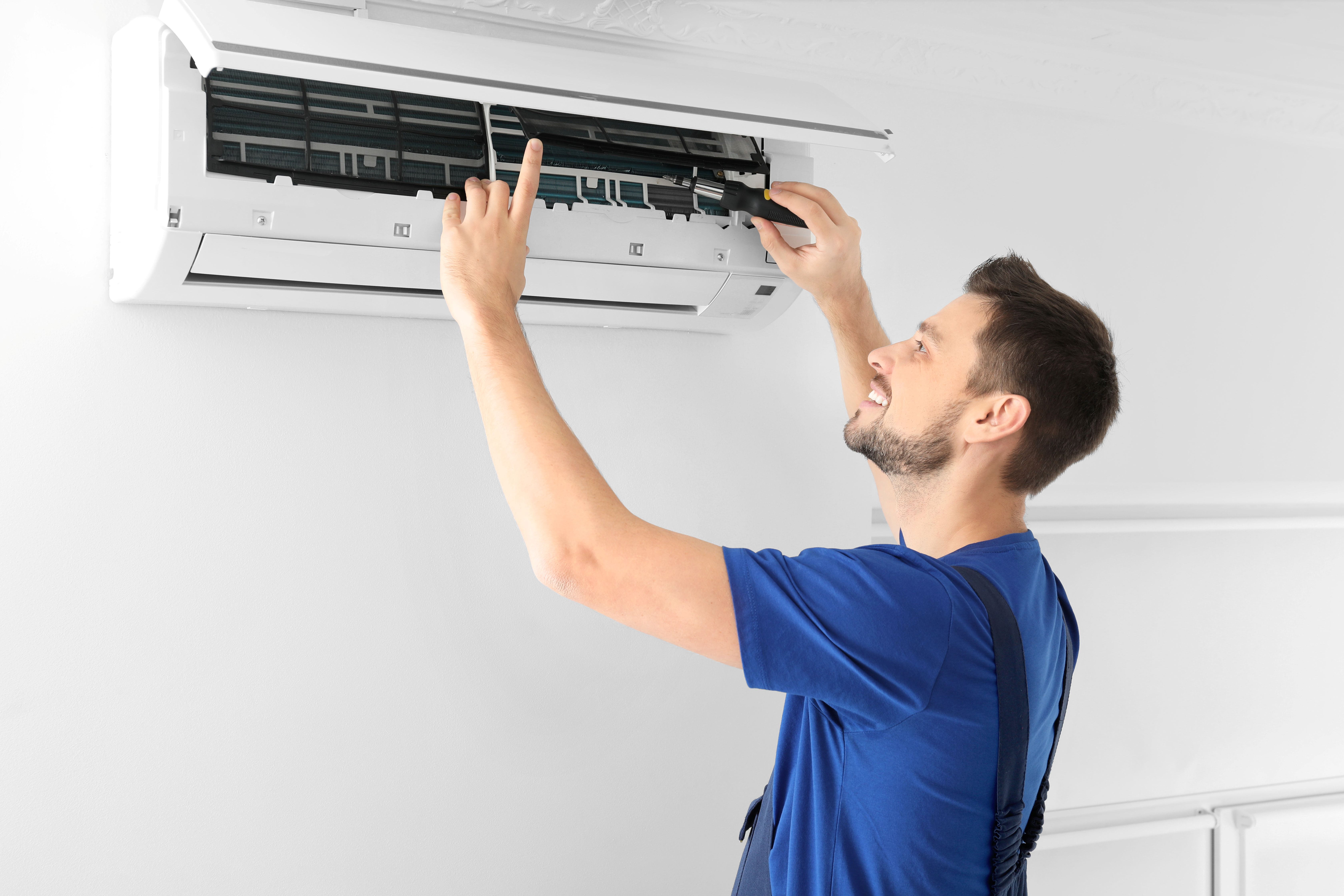 Technician wearing gloves and a blue cap installing or servicing an outdoor air conditioning unit on a white wall.