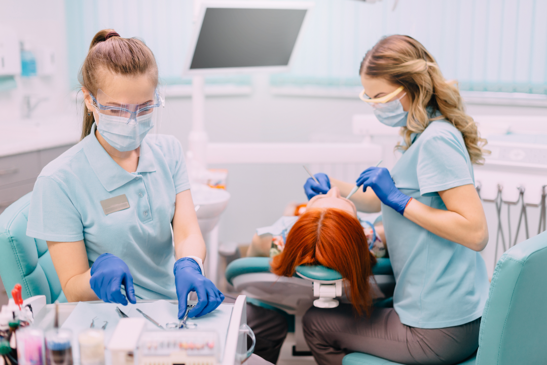 Two female dental professionals wearing masks, gloves, and protective eyewear working on a patient's teeth in a modern dental clinic.