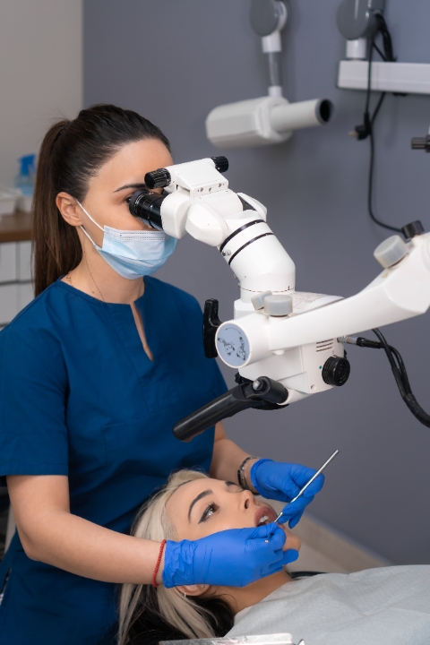Female dentist in blue scrubs and mask using a dental microscope to examine a patient's mouth in a dental clinic.