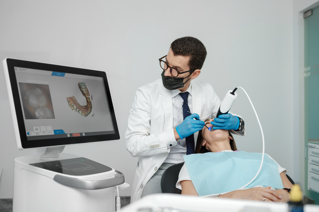 Dentist wearing gloves and mask using a digital scanner on a female patient’s teeth with a 3D dental image displayed on a monitor.