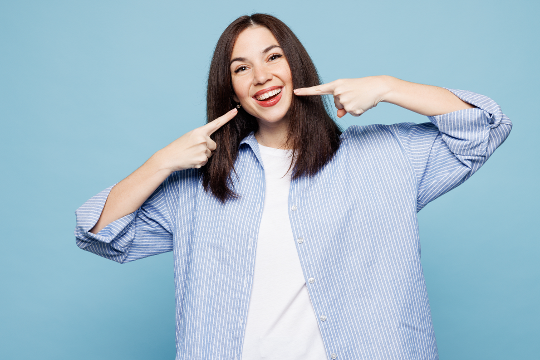Smiling young woman wearing a blue striped shirt pointing with both index fingers to her teeth against a blue background.
