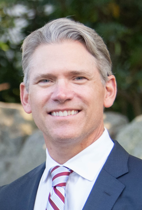 Smiling middle-aged man with gray hair wearing a dark suit, white shirt, and striped tie.