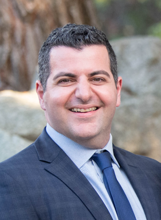 Smiling man with short dark hair wearing a blue suit, light blue shirt, and navy tie standing outdoors with blurred trees and rocks in the background.