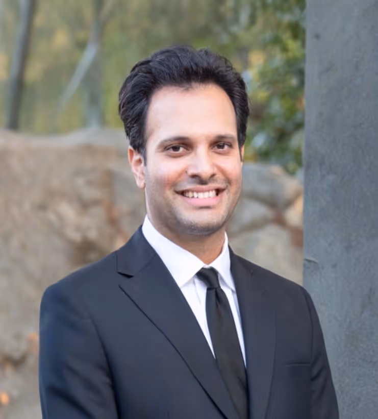 Smiling man with dark hair wearing a black suit and tie standing outdoors near a stone and greenery.