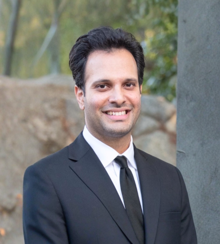 Smiling man with dark hair wearing a black suit and tie standing outdoors near a stone and greenery.