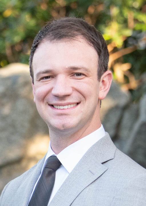 Smiling man wearing a light gray suit jacket, white shirt, and black tie standing outdoors with trees and rocks in the background.