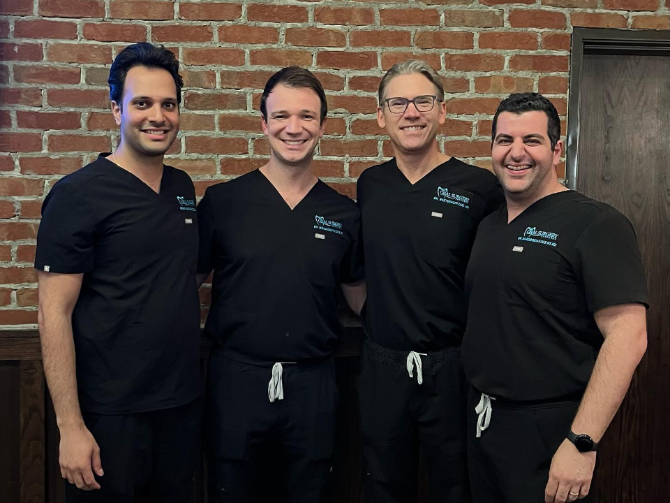 Four men wearing black medical scrubs standing and smiling in front of a brick wall.