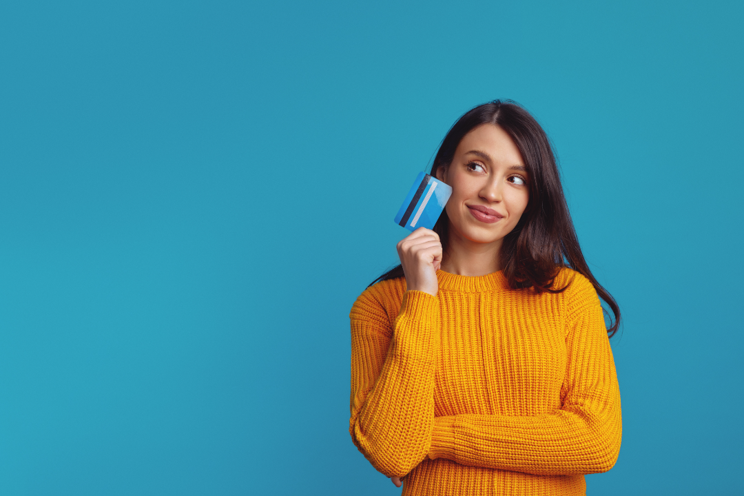 Young woman in an orange sweater holding a credit card and looking to the side against a blue background.