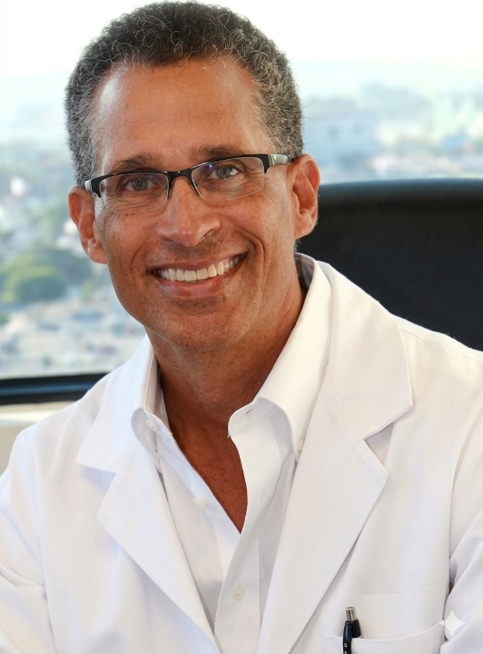 Smiling man with gray curly hair and glasses wearing a white lab coat, sitting indoors with a city view in the background.