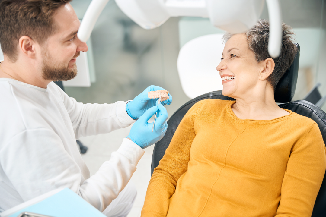 Dentist wearing blue gloves showing a dental mold to a smiling middle-aged woman in a mustard sweater sitting in a dental chair.