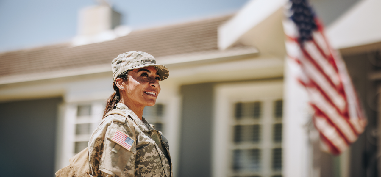 Smiling female soldier in camouflage uniform with American flag patch standing outside a house with a blurred American flag in the background.