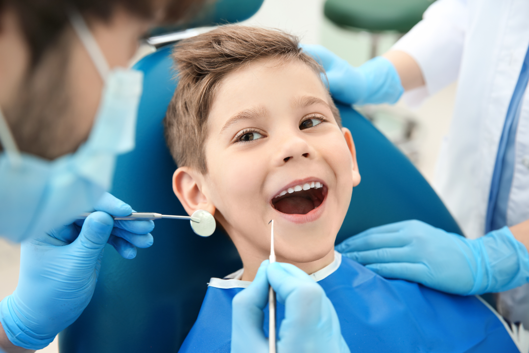 Smiling boy sitting in a dental chair with mouth open while a dentist in blue gloves examines his teeth using dental tools.