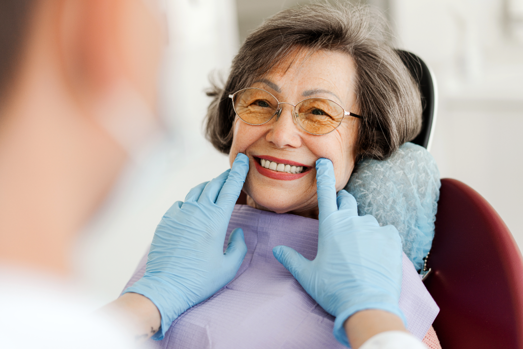 Smiling elderly woman wearing glasses at the dentist with gloved hands gently holding her cheeks.