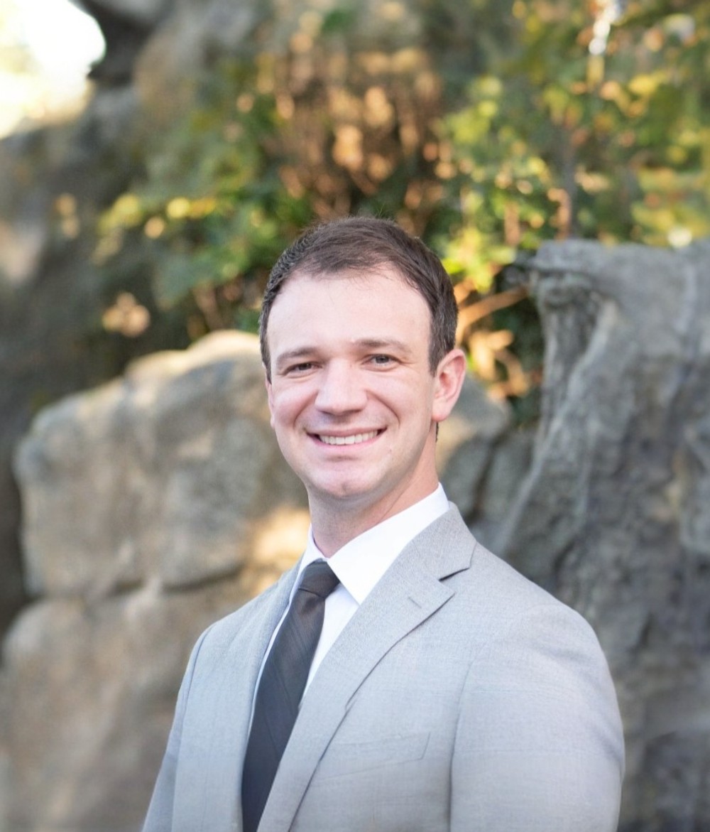 Smiling man in a light gray suit and black tie standing outdoors with rocks and greenery in the background.