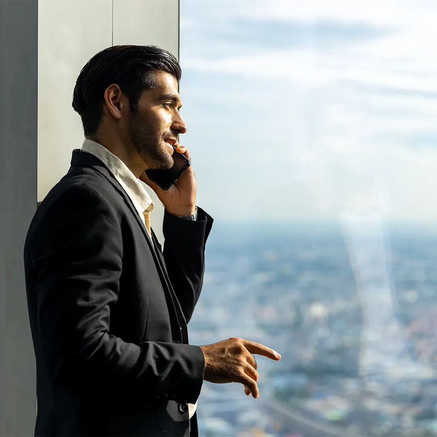 Man in a black suit talking on a smartphone while standing by a window with a cityscape view.