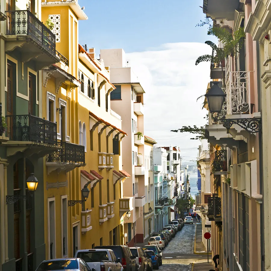 Narrow street with parked cars and colorful buildings under clear sky leading to a distant view of the ocean.