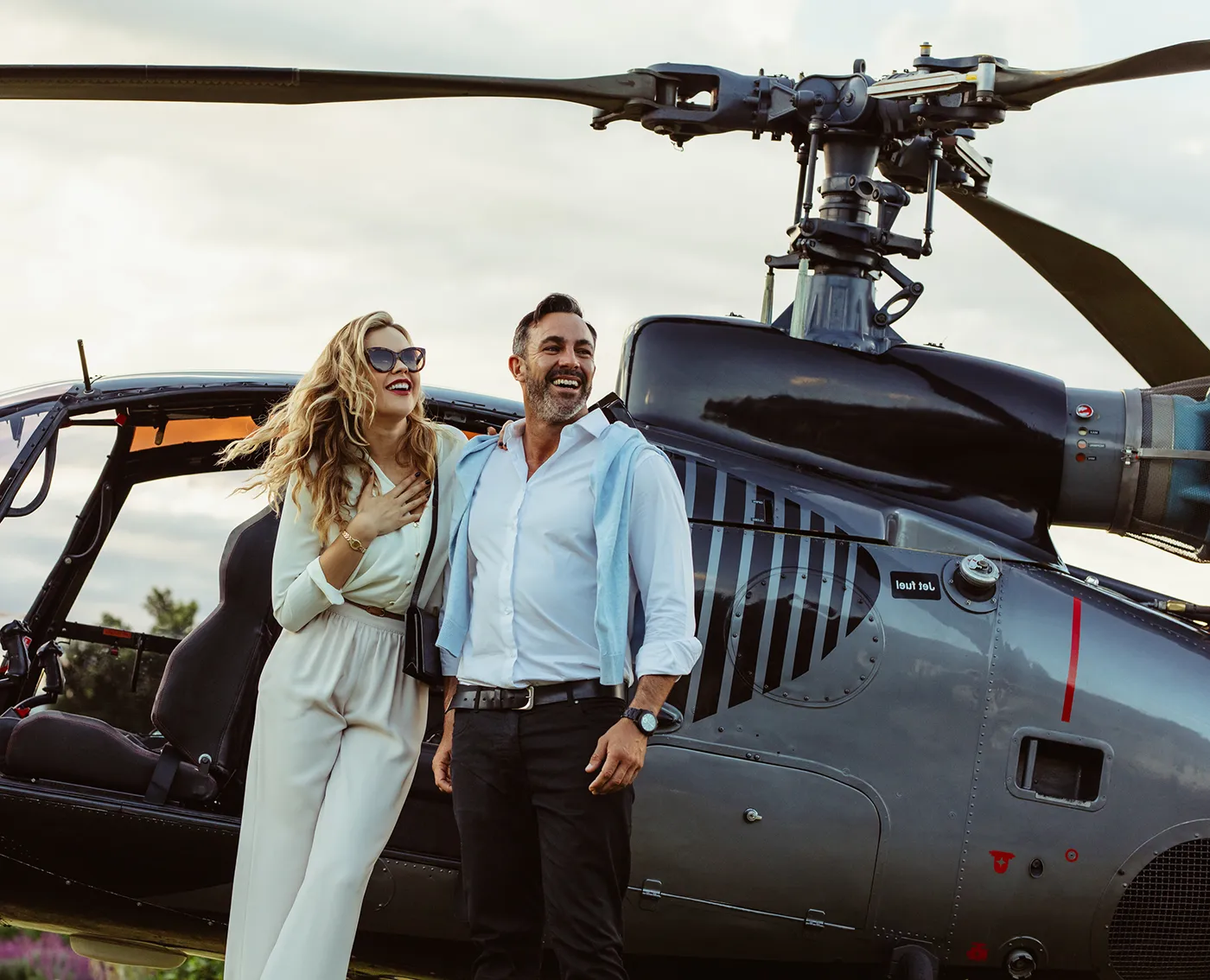 Smiling couple standing together beside a gray helicopter with the woman wearing sunglasses and white outfit.