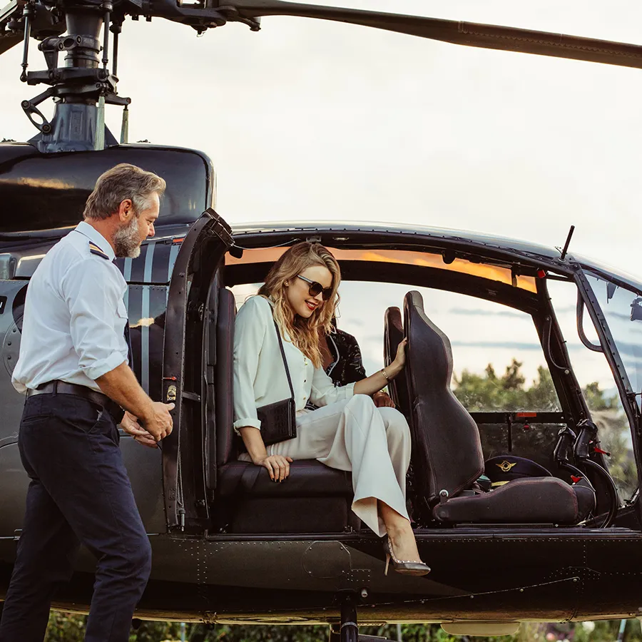 Woman in white outfit and sunglasses sitting inside a black helicopter with pilot standing outside near the open door.