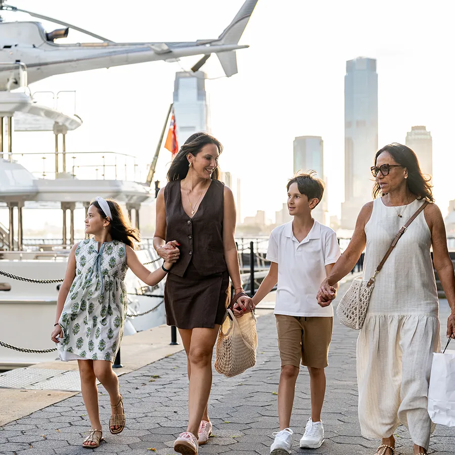Four people walking hand in hand on a waterfront promenade with yachts and city skyscrapers in the background.