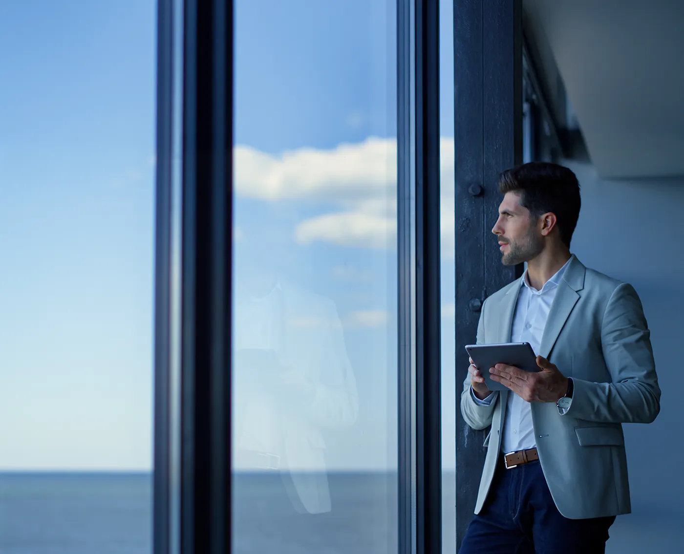 Man in a light gray blazer holding a tablet and looking out a large window at a blue sky and ocean.