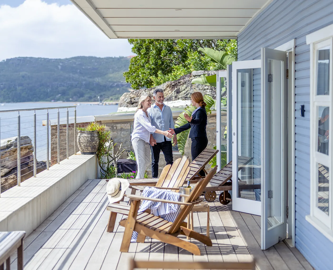 Couple greeting a real estate agent on a wooden deck with lounge chairs overlooking water and hills.