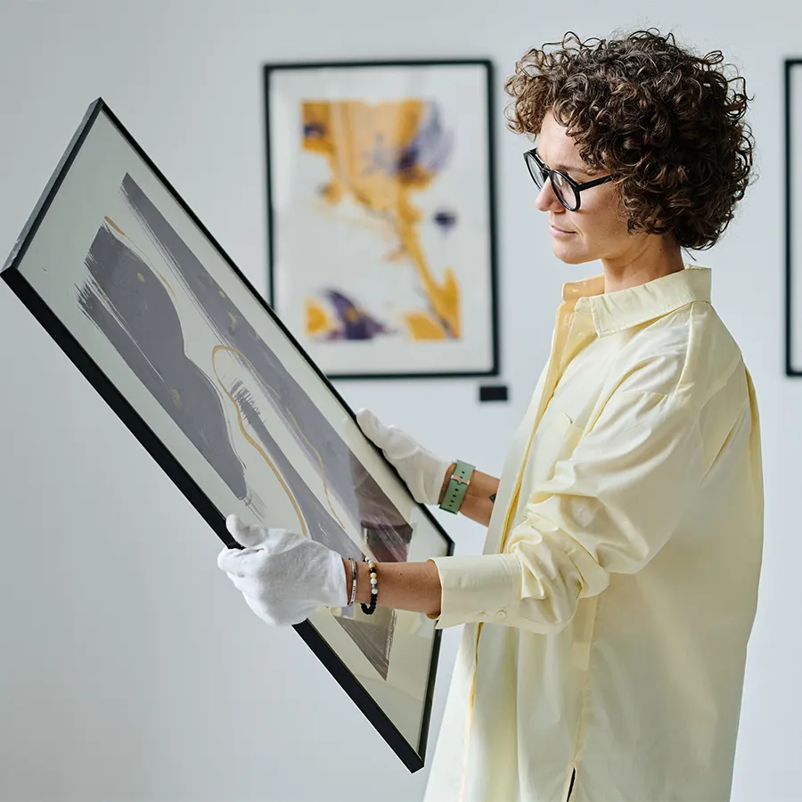 Woman wearing white gloves examining a framed abstract artwork in a gallery.