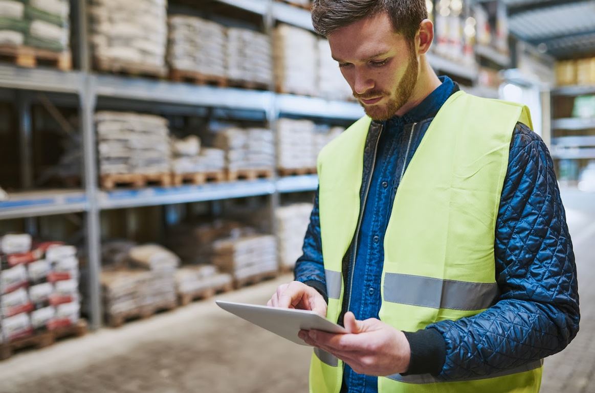 Health and safety consultant in a hi-vis vest using a tablet to record findings during a gap analysis walkthrough at a builders merchant warehouse.