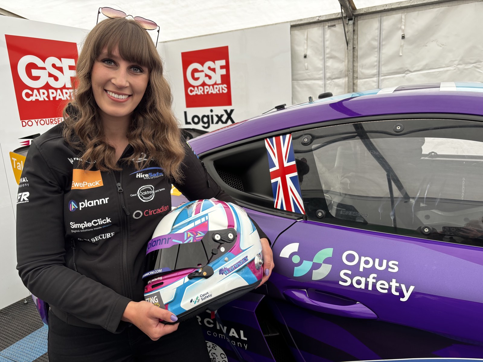 Lydia Walmsley holding her racing helmet beside her Porsche race car, both carrying Opus Safety branding