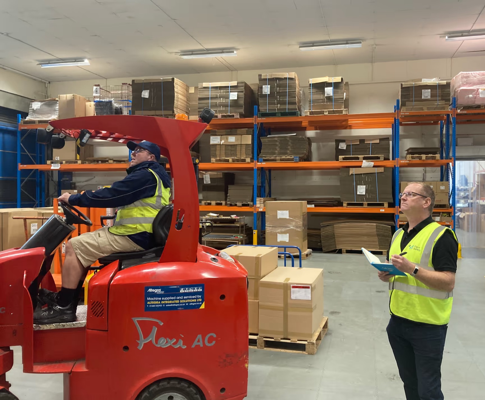 An Opus Safety consultant in hi-vis with a notepad, watching a forklift operator in a warehouse with pallet racking