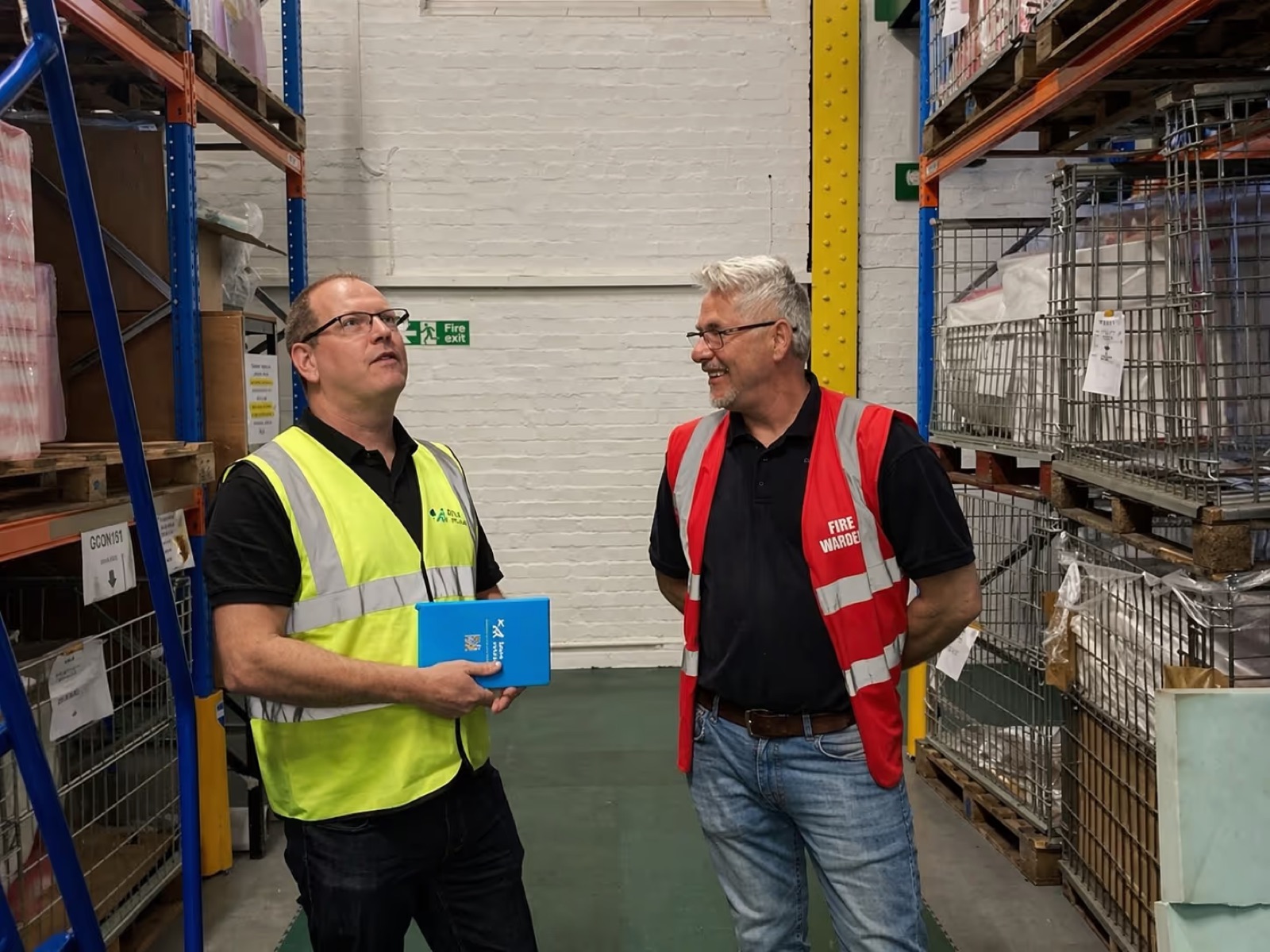 Health and safety consultant in a hi-vis vest discussing a safety guide with a fire warden in a warehouse
