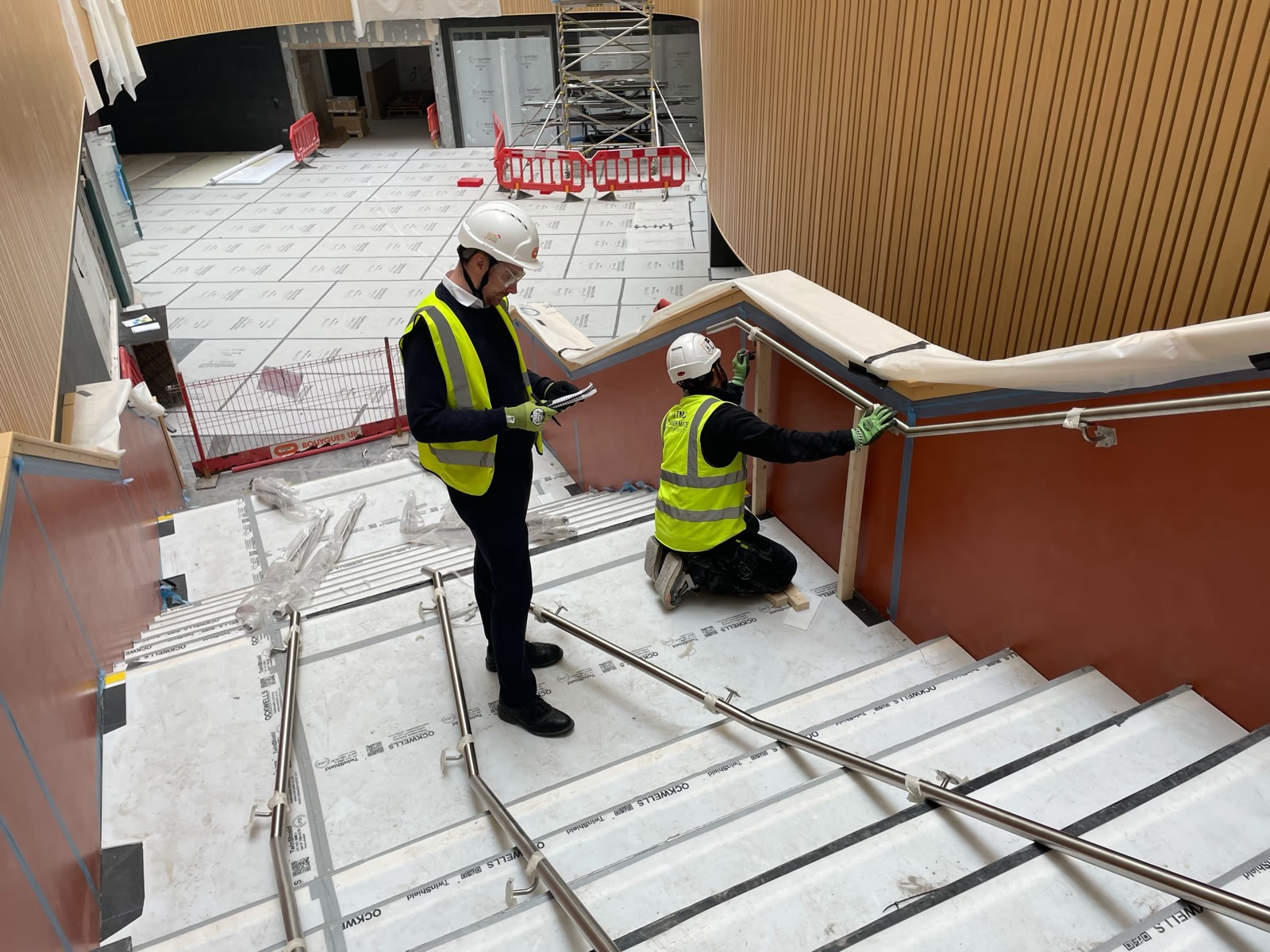 Health and safety consultant watching a handrail being fitted on a construction staircase, with a scaffold tower behind