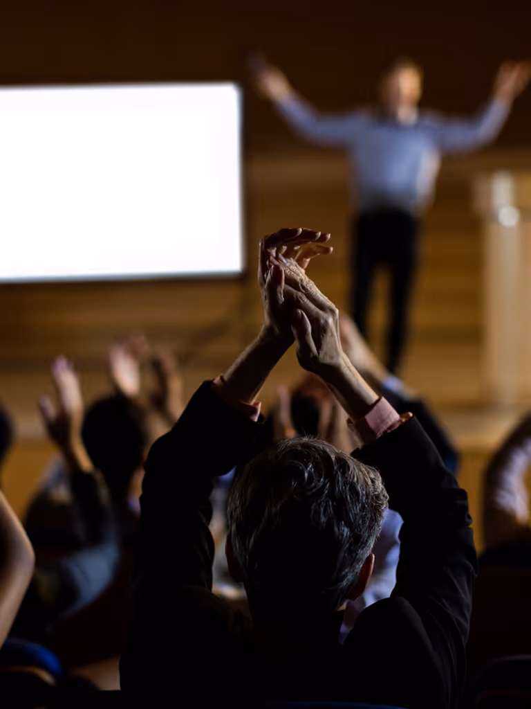 Audience clapping with hands raised in front of a presenter on a stage with a bright screen in the background.