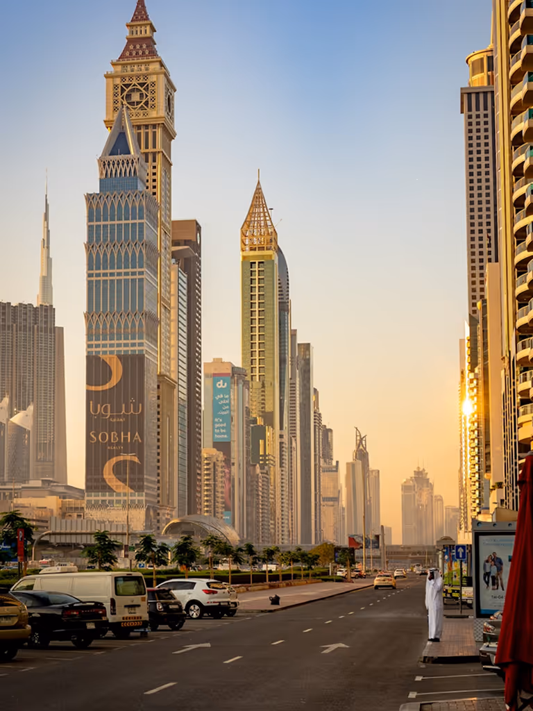 Wide city street in Dubai with high-rise buildings, including the Burj Khalifa in the background at sunset.