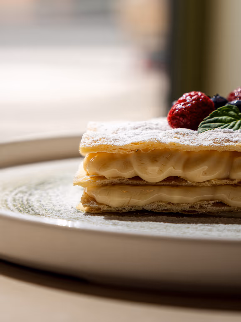 Cream-filled layered pastry dusted with powdered sugar, topped with raspberries and a mint leaf on a white plate.