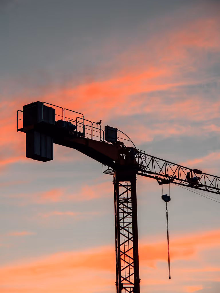 Silhouette of a construction crane against a colorful sunset sky with orange and pink clouds.