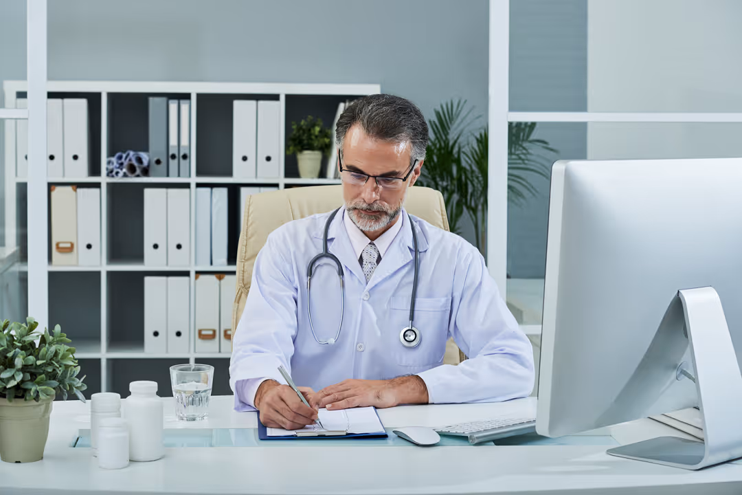 Male doctor with glasses and stethoscope writing on a clipboard at a desk in a modern office.